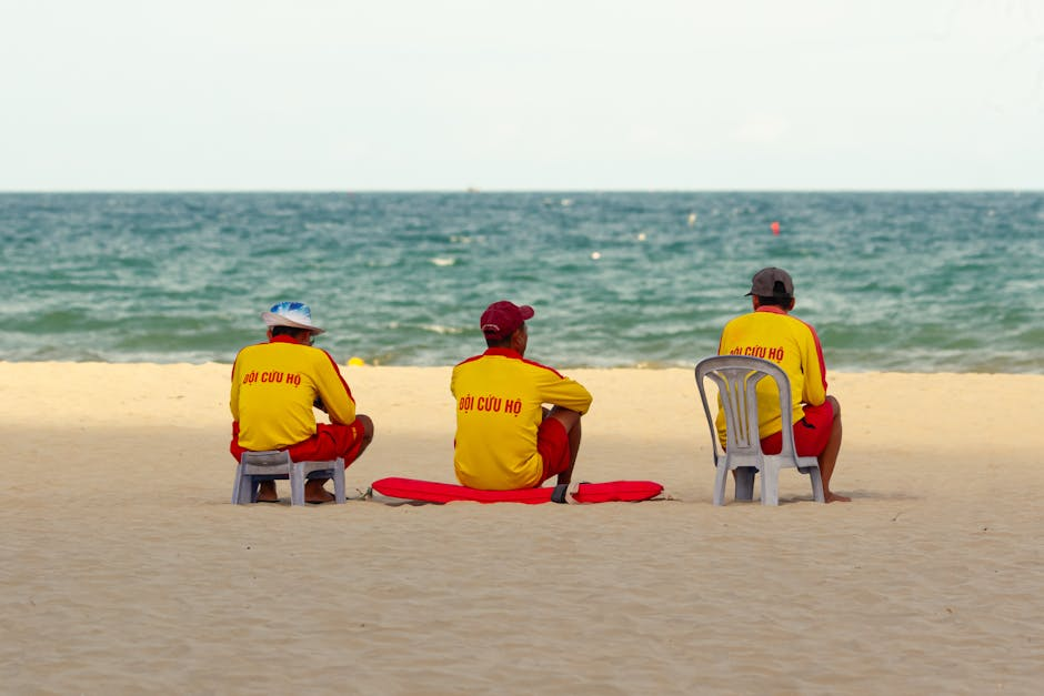 Three lifeguards in red and yellow uniforms sitting on a sandy beach facing the sea.